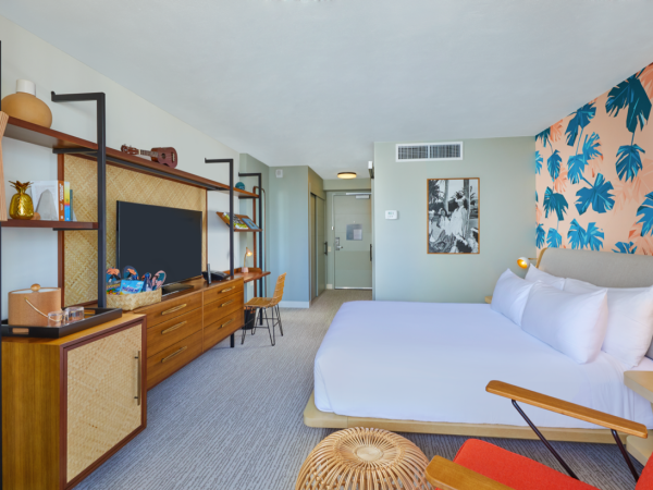A stylish hotel room with a large bed, vibrant leaf-patterned wallpaper, a TV, and mid-century modern furnishings.