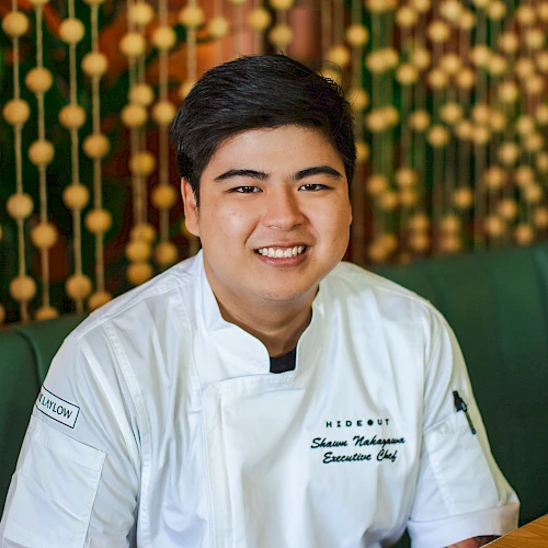 A smiling person wearing a chef's uniform with tattooed arm, sitting in a restaurant with a decorative background.