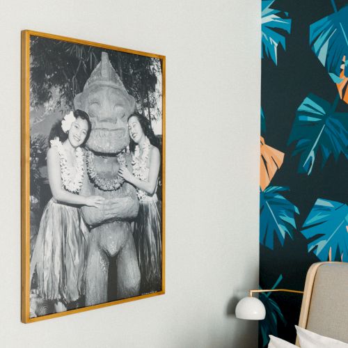 The image shows a vintage-style black and white photo of two women in hula skirts with a statue, hanging on a wall with tropical wallpaper.