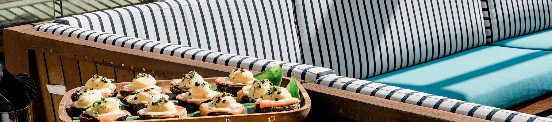 Outdoor buffet on a terrace with sliders, shrimp skewers, and salads beside striped loungers and greenery.