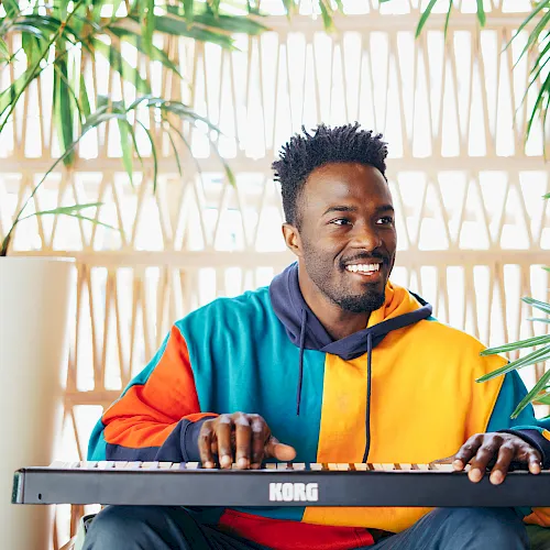 A smiling man in a colorful hoodie plays a KORO keyboard by bright windows, with green plants nearby.