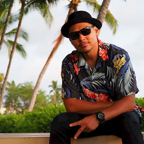 A man in a black hat and sunglasses, wearing a floral shirt, relaxing by palm trees in a tropical outdoor setting.