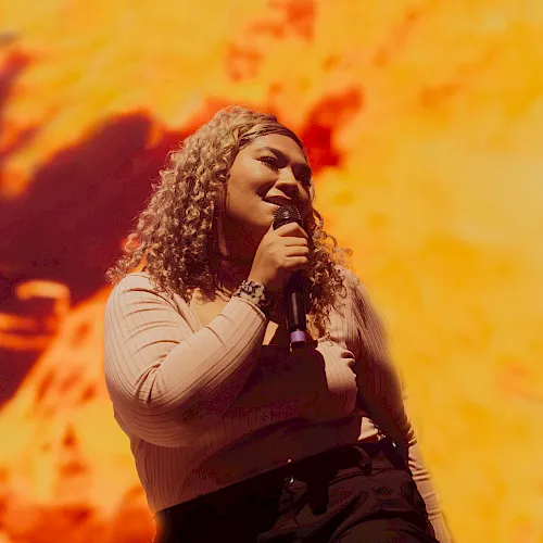A woman with curly hair sings into a microphone on stage under warm orange concert lights, smiling as she performs.