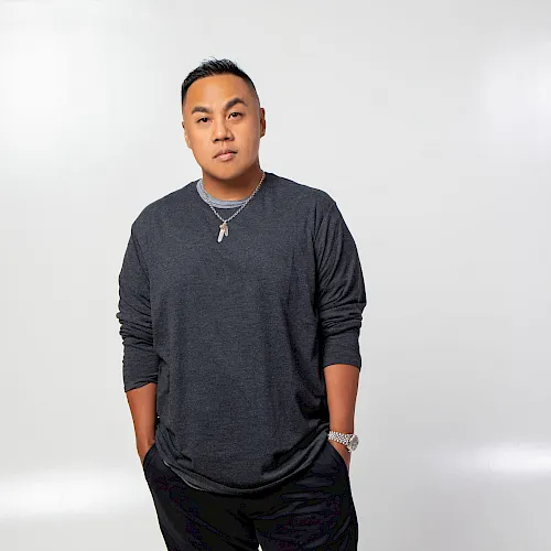 A man poses in a studio against a plain white background, wearing a dark long-sleeve shirt, necklace, and watch.
