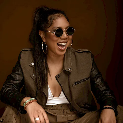 Smiling woman wearing round sunglasses and hoop earrings in a studio portrait with a leather jacket against a brown backdrop.