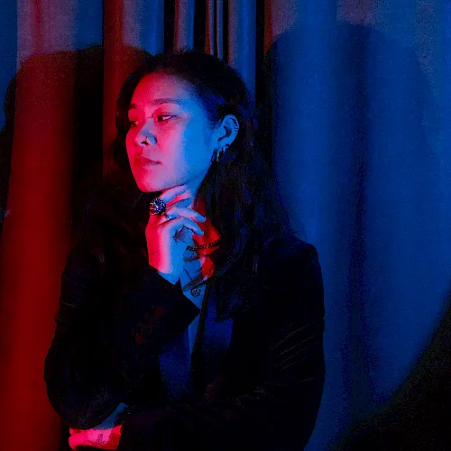 A woman in a dark suit poses with red-blue stage lighting, hand near her chin, standing before dark curtains.