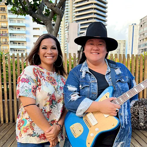 Two smiling women pose outdoors; one holds a blue electric guitar on a patio with city apartments behind them.