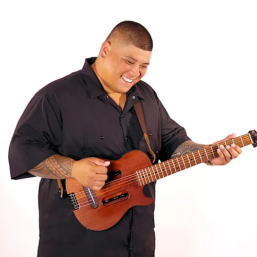 A smiling man plays a brown electric guitar, leaning forward with his fingers on the frets.