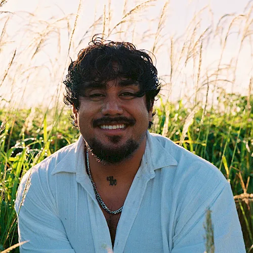A smiling man sits in a sunlit field of tall grass, looking at the camera.