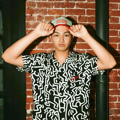 A teen in a patterned button-up and cap marked “500N” poses with both hands on his hat against a brick wall, relaxed.
