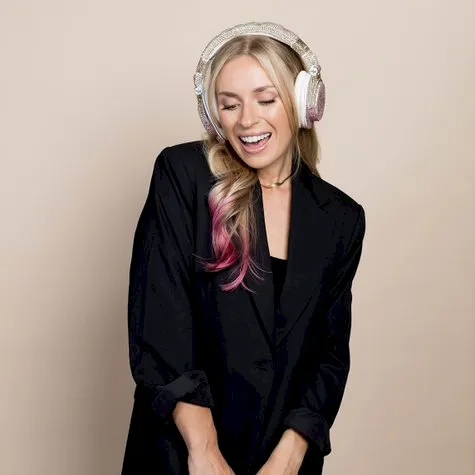 A smiling woman in a black blazer dress wearing over-ear headphones stands against a plain beige background with her hands clasped.