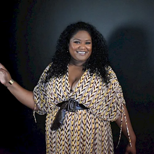 A woman with long curly hair in a patterned yellow-and-white dress smiles and gestures beside a dark studio backdrop.