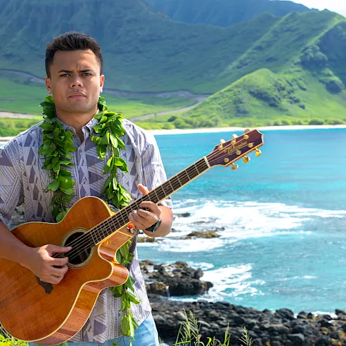 A man strums an acoustic guitar with a green lei at a rocky beach, with turquoise ocean and green mountains behind him.