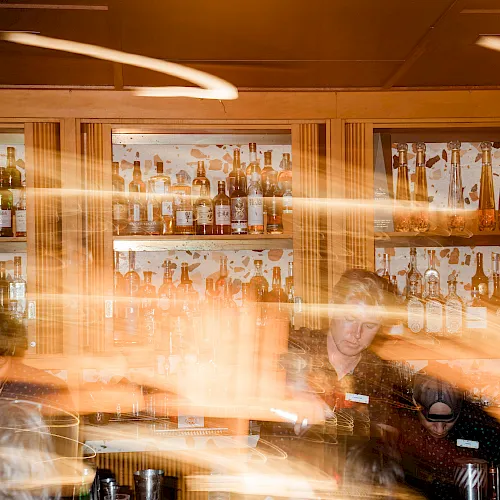 A bar liquor shelf packed with bottles, warm lights, and a bartender with motion blur in the foreground behind the glass.