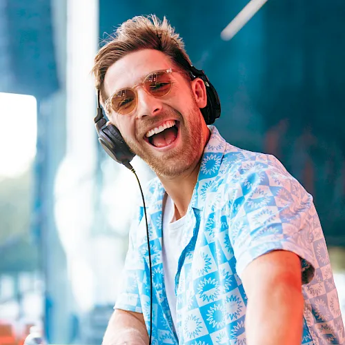 A smiling DJ wearing over-ear headphones and mirrored sunglasses, grinning into a microphone during a lively performance.