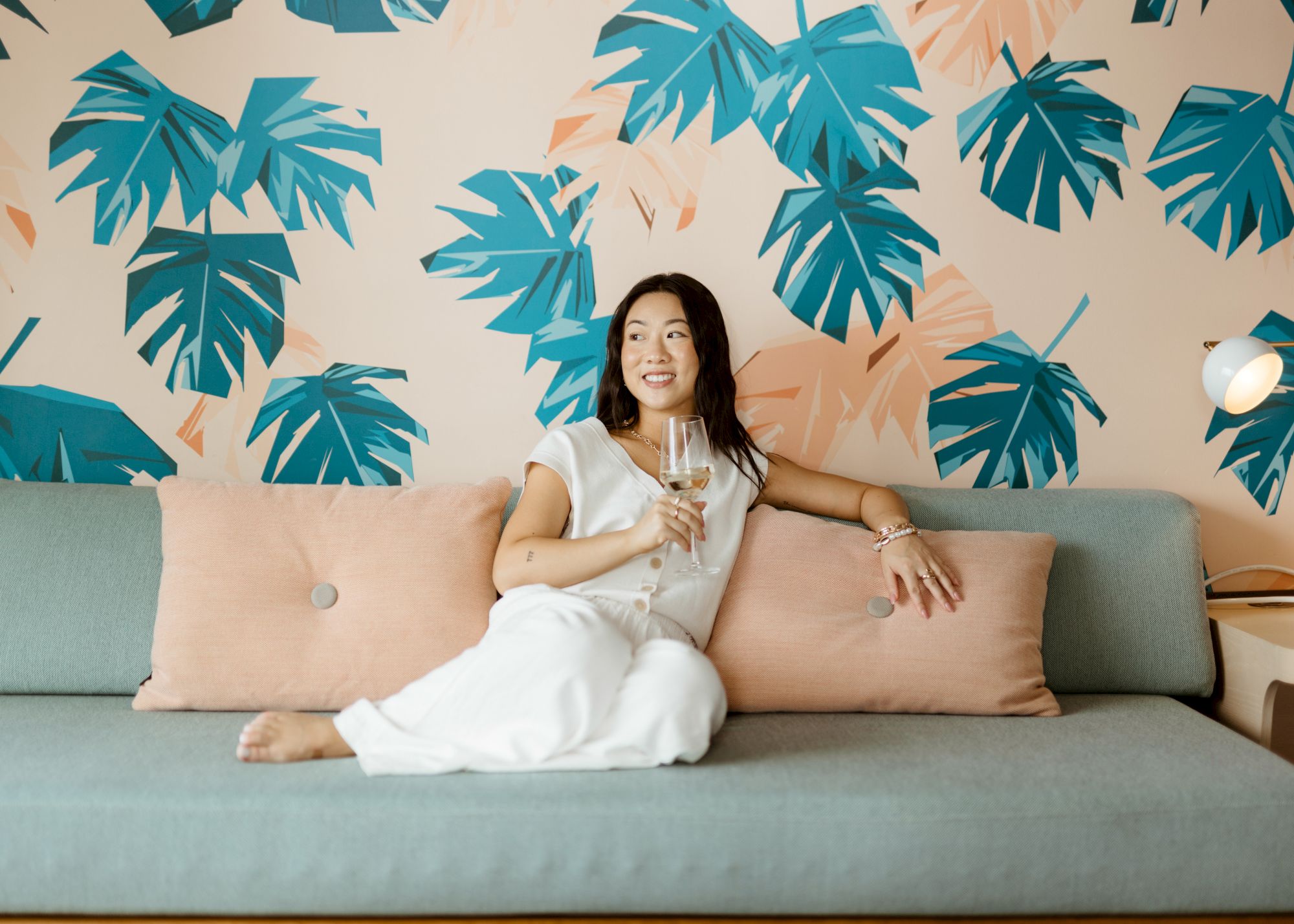 A smiling woman in a white dress sits on a light gray couch with peach cushions, tropical teal palm leaf wallpaper, and a small lamp nearby.