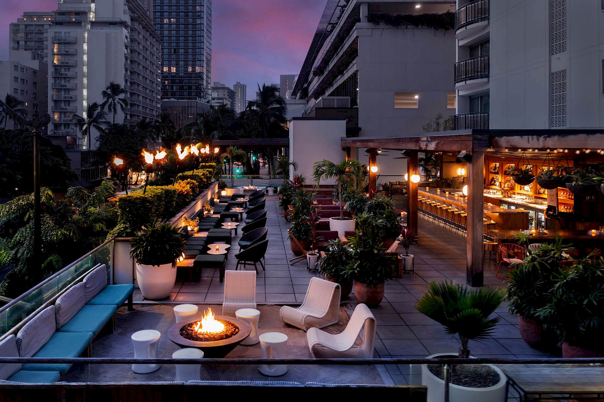 The image shows a rooftop bar with seating, a fire pit, and ambient lighting, surrounded by city buildings, under a twilight sky.