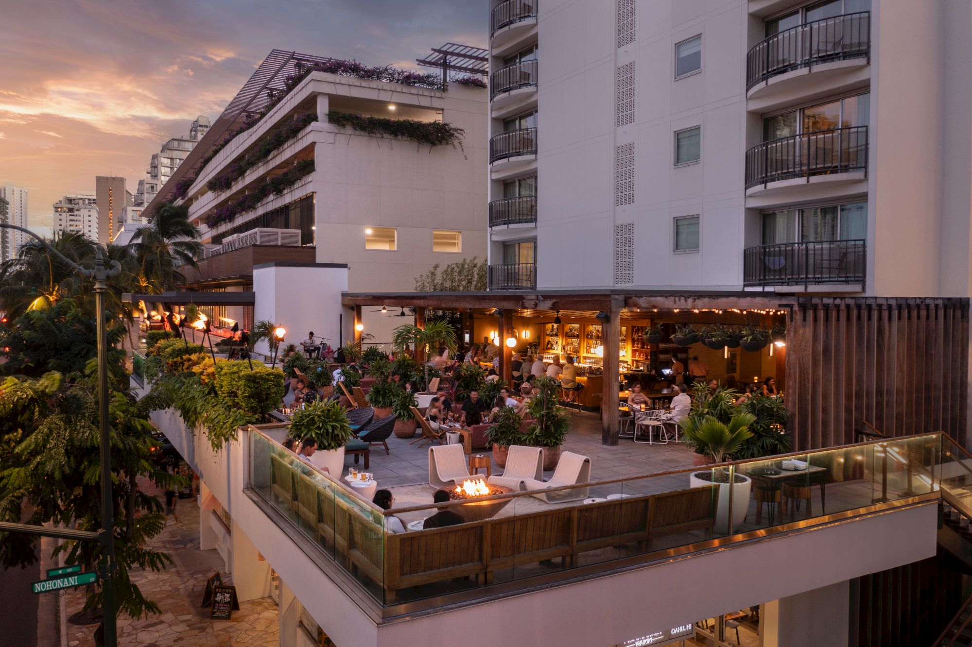 A seaside hotel terrace at dusk with people dining, warm string lights, outdoor lounge chairs, potted plants, and a modern white building backdrop.