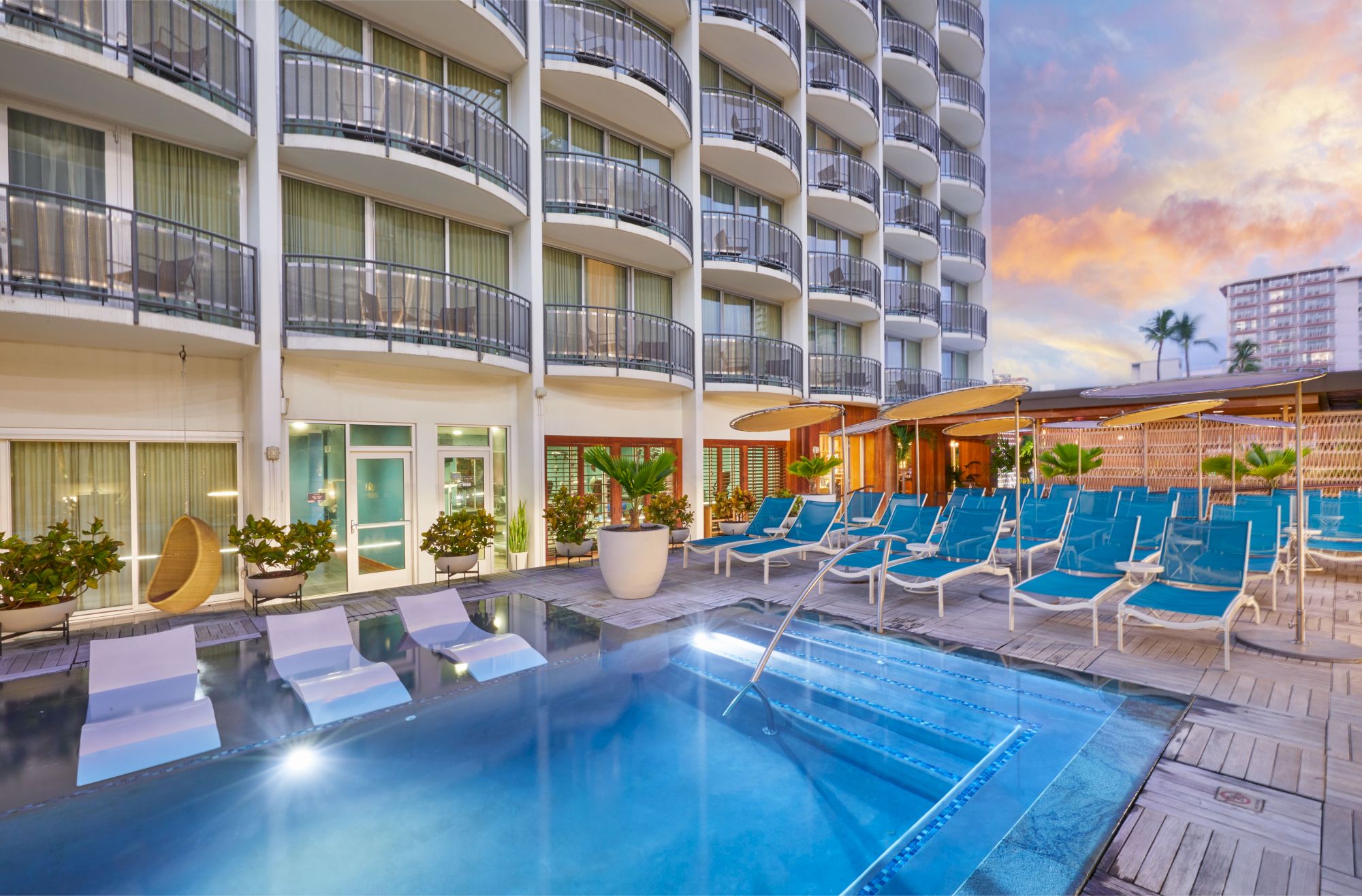 A resort-style pool area with blue lounge chairs, umbrellas, and a multi-story hotel building in the background; sunset sky overhead.
