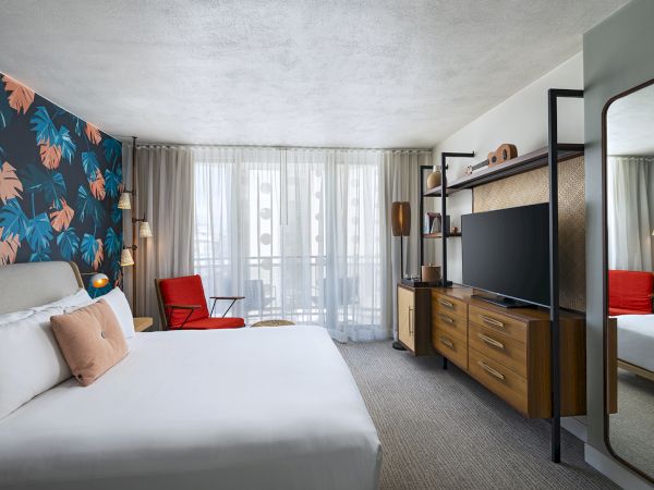 A modern hotel room with a patterned feature wall, white bed, red chair, large window, and a wooden dresser with a flat-screen TV; full-length mirror nearby.