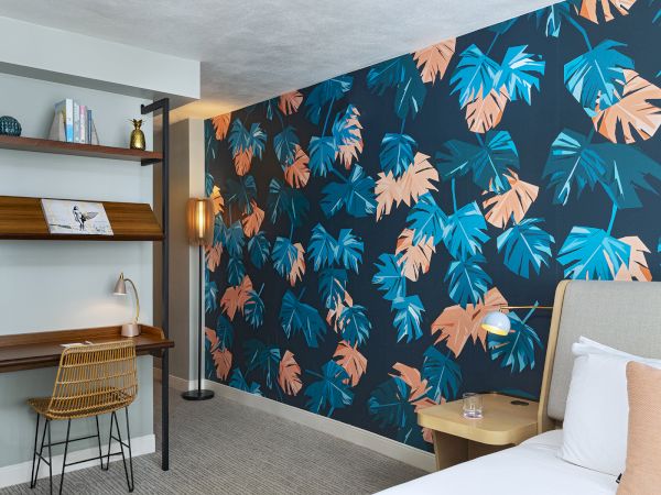 A modern bedroom with a tropical leaf wallpaper, a small desk and chair, a shelving unit, and a neatly made bed with peach accents.