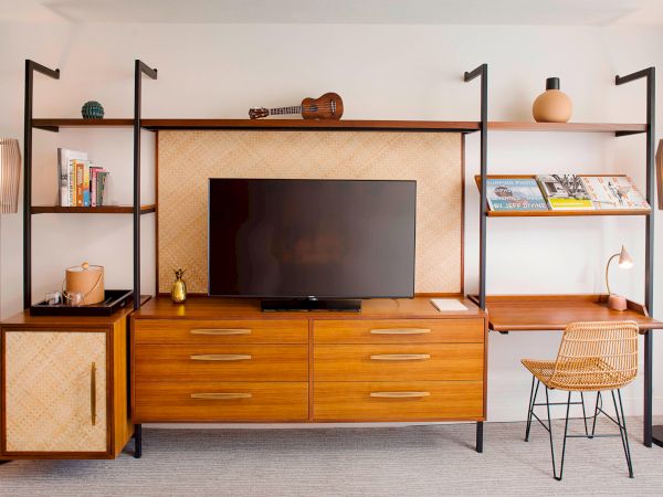 A stylish wooden entertainment unit with a TV, books, magazines, a small guitar, decorative items, and a desk with a chair.