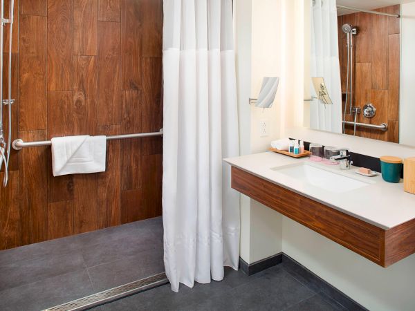 The image shows a modern bathroom with a walk-in shower, white curtain, wooden accents, sink, and mirror.