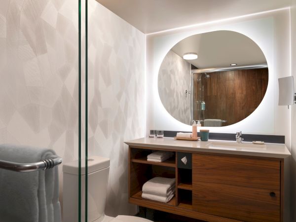 The image shows a modern bathroom with a round mirror, a wooden vanity, and towels. A shower and a toilet are also visible in the space.
