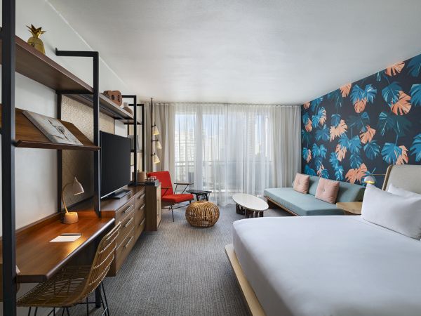 A stylish hotel room with a tropical floral accent wall, white bed, seating area, desk, and a large window letting in natural light.