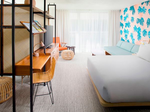 A modern hotel room with a bed, desk, chair, and a couch. The wall has a tropical leaf design. Light streams through sheer curtains.
