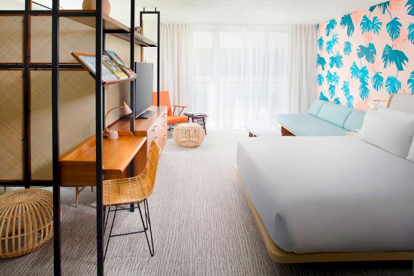 A modern hotel room with a bed, desk, chair, and a couch. The wall has a tropical leaf design. Light streams through sheer curtains.