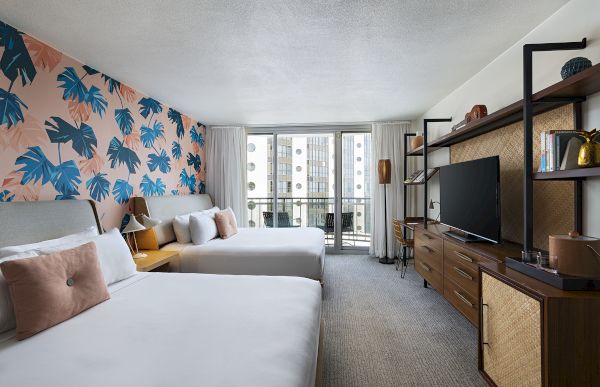A bright hotel room with two beds, tropical leaf wallpaper, a wall-mounted TV, wooden dresser, and a balcony view of city buildings, stylish and airy.