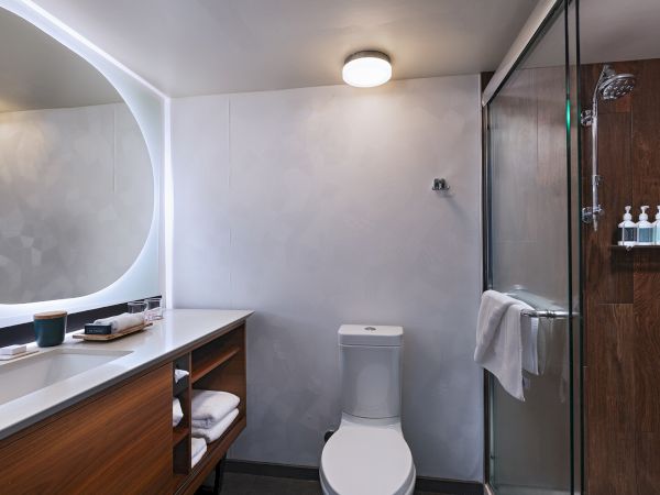 A modern bathroom with a rounded backlit mirror, wooden vanity, open shelving, a toilet, and a glass-enclosed shower with a towel bar and toiletries, ending with a period.
