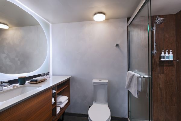 A modern bathroom with a rounded backlit mirror, wooden vanity, open shelving, a toilet, and a glass-enclosed shower with a towel bar and toiletries, ending with a period.