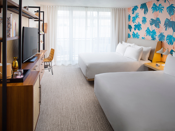 A hotel room featuring two beds, a tropical-themed accent wall, a TV on a stand, a desk with a chair, and large windows with sheer curtains.