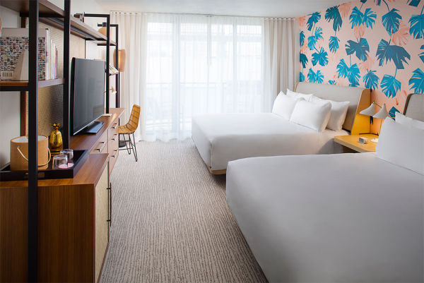 A bright hotel room with two white beds, a patterned blue tropical wallpaper accent wall, a desk, TV, and large windows with sheer curtains. Top it at 140 characters, always ending the sentence.