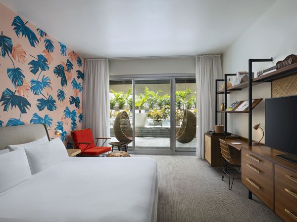 A modern hotel room with a palm wallpaper accent wall, a comfy bed, a red chair, a balcony with hanging chairs, and a full wooden dresser and desk setup.