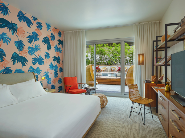 The image shows a modern bedroom with tropical-themed wallpaper, a bed, a TV, and a view of an outdoor patio through sliding doors.