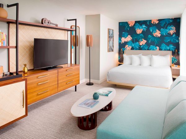 A modern hotel room with a bed, TV on a wooden cabinet, sofa, and a colorful tropical-themed accent wall.