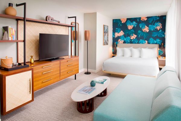 A modern hotel room with a bed, TV on a wooden cabinet, sofa, and a colorful tropical-themed accent wall.