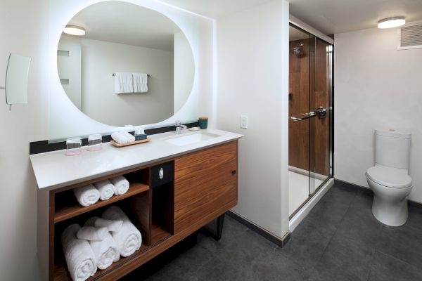 A modern bathroom with a circular mirror, wooden vanity with towels, glass shower, and toilet on grey-tiled floor.