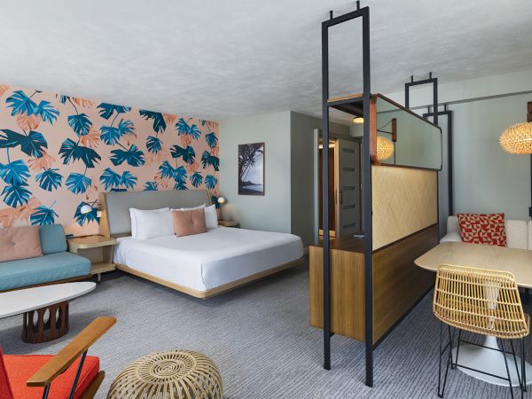 A modern hotel room with a bed, seating area, and dining table. Decor features a bold leaf-patterned wall and warm lighting accents.