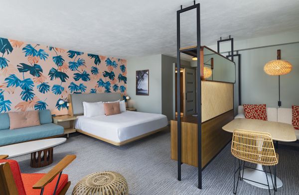 A modern hotel room with a bed, seating area, and dining table. Decor features a bold leaf-patterned wall and warm lighting accents.