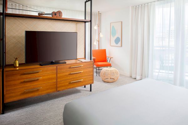A modern hotel room with a wooden TV stand, an orange chair, and a bed. Light color scheme enhances spaciousness and elegance.