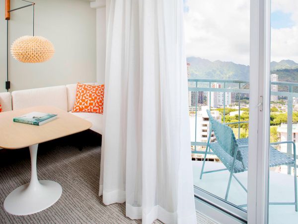 A cozy room with a table, white curtains, and a view of city buildings and mountains through the window.