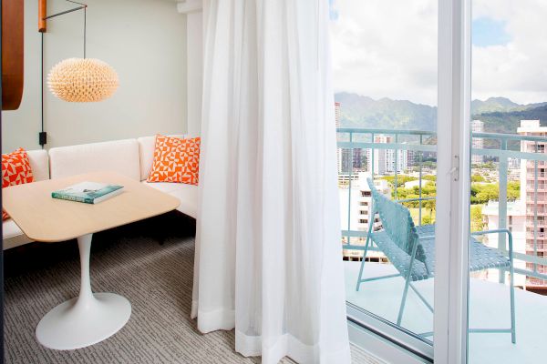 A cozy room with a table, white curtains, and a view of city buildings and mountains through the window.