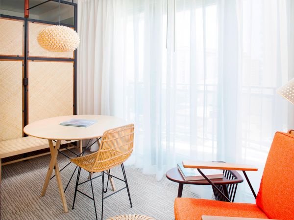 The image shows a modern room with an orange armchair, round table, wicker chair, and a woven pendant light near sheer curtains.
