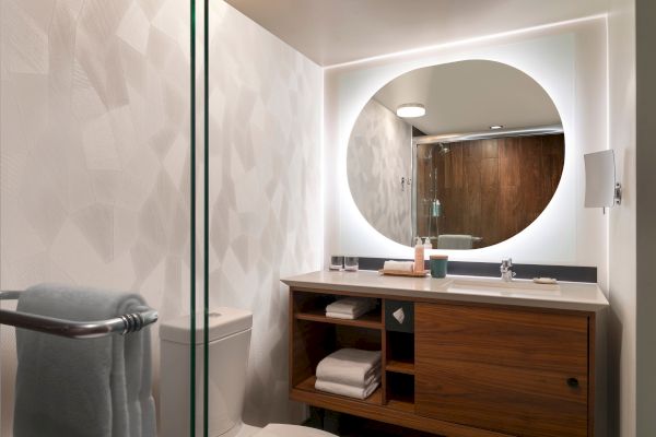 The image shows a modern bathroom with a round mirror, wooden vanity, towels, and a toilet, featuring sleek, minimalist design elements.