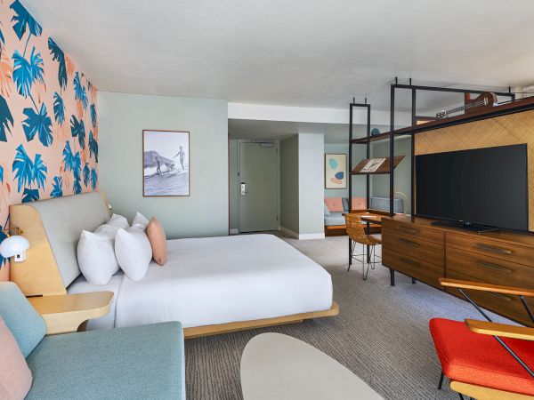 A modern hotel room with a bed, TV, desk, and colorful decor featuring a leaf-patterned wall and sleek furniture.