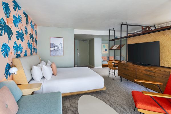 A modern hotel room with a bed, TV, desk, and colorful decor featuring a leaf-patterned wall and sleek furniture.